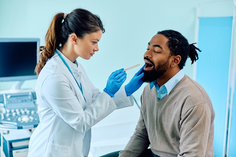 Female doctor using tongue depressor while checking black man's throat at medical clinic.