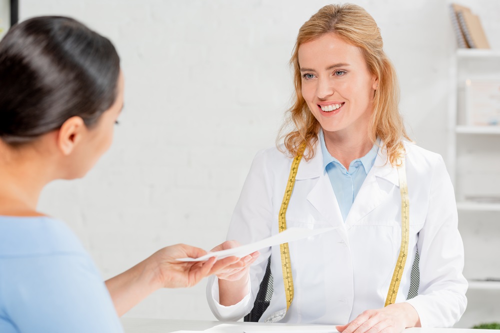 attractive and smiling nutritionist sitting at table and giving paper to patient in clinic